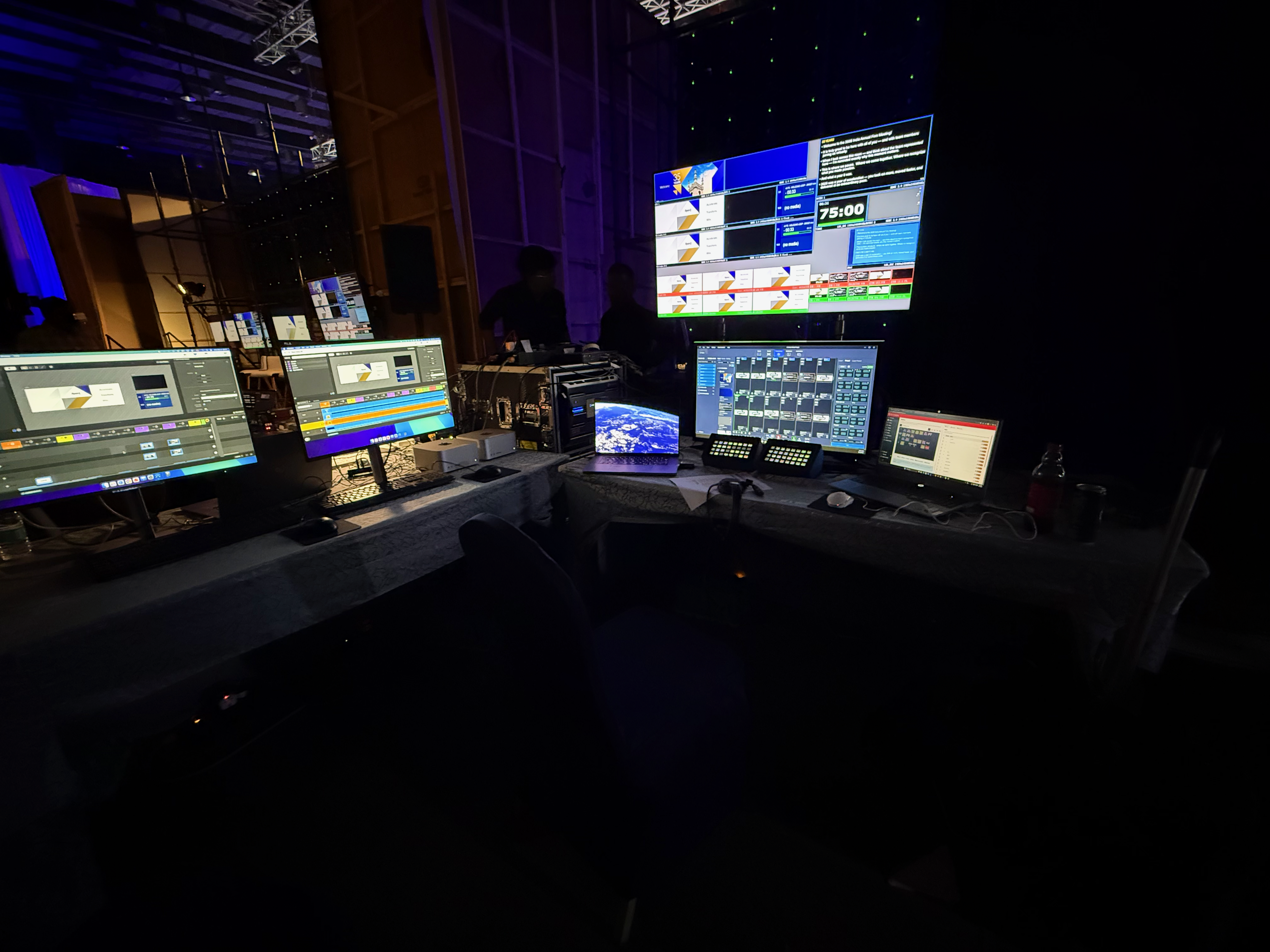 Dark control room with glowing monitors displaying video editing software and live broadcast feeds.