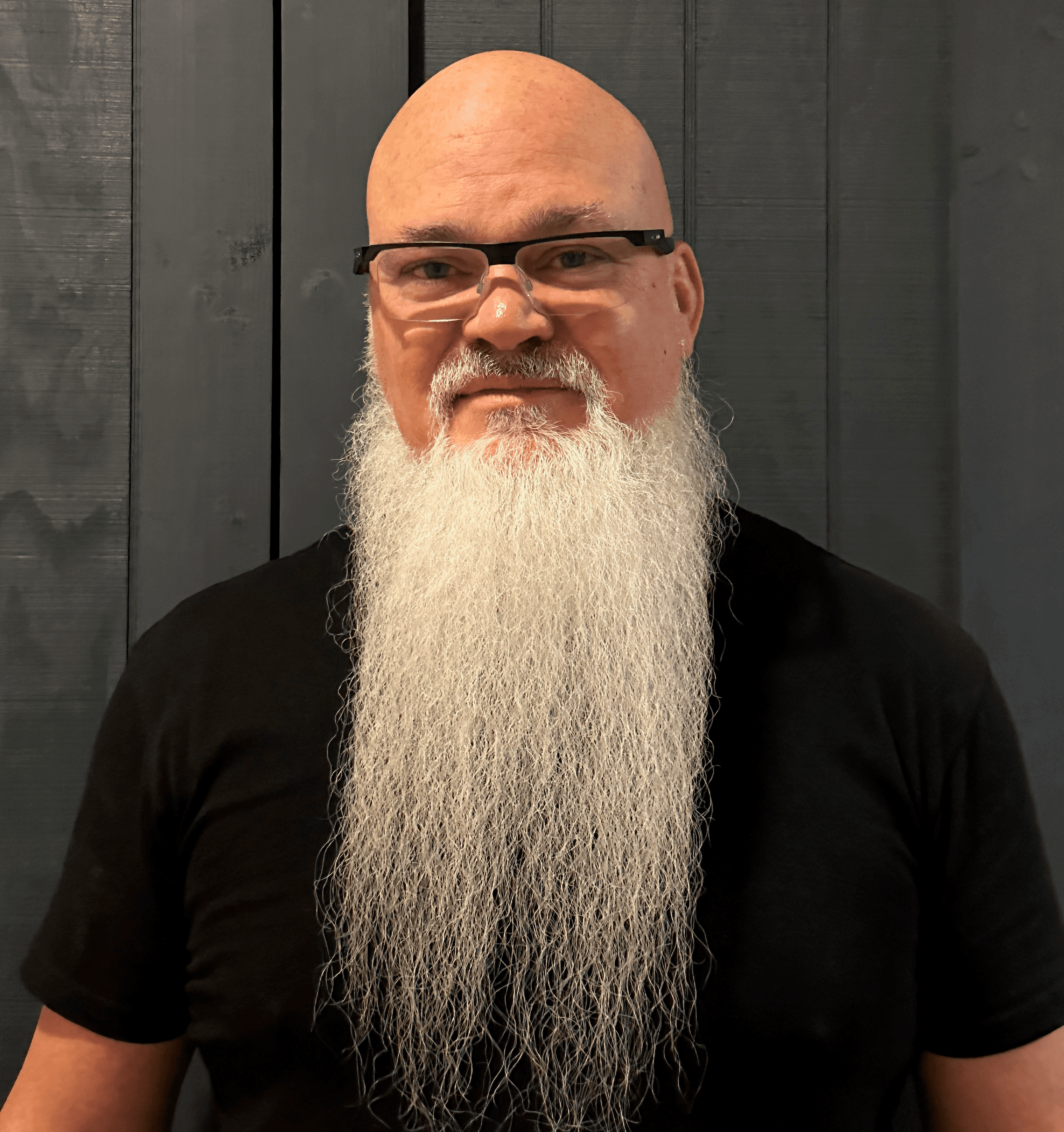 Bald man with glasses and a long, flowing white beard against dark wooden paneling.